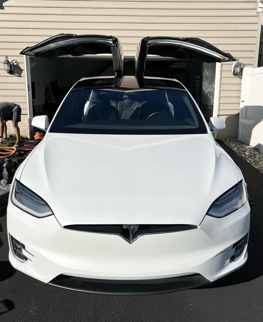 White Tesla with open falcon-wing doors parked in a driveway, front view.