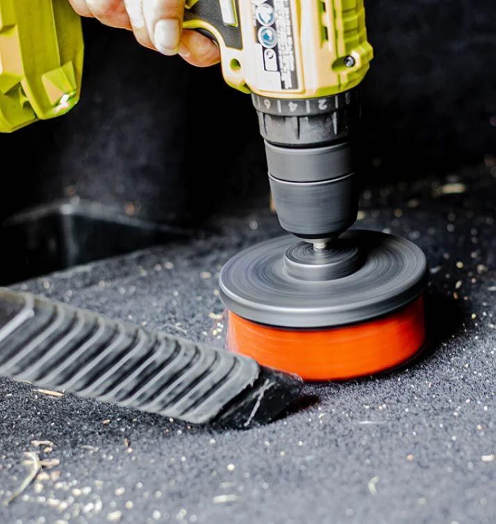 A power drill with a sanding attachment smoothing a black surface beside a broom brush.