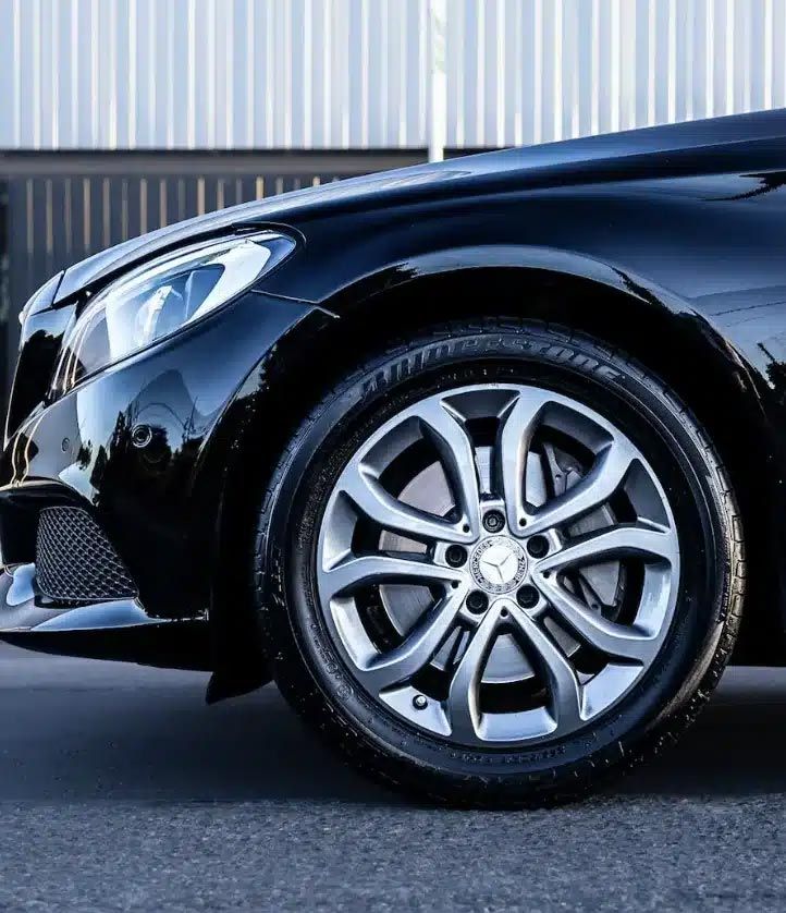 Close-up of a black car’s front wheel and headlight parked on pavement.