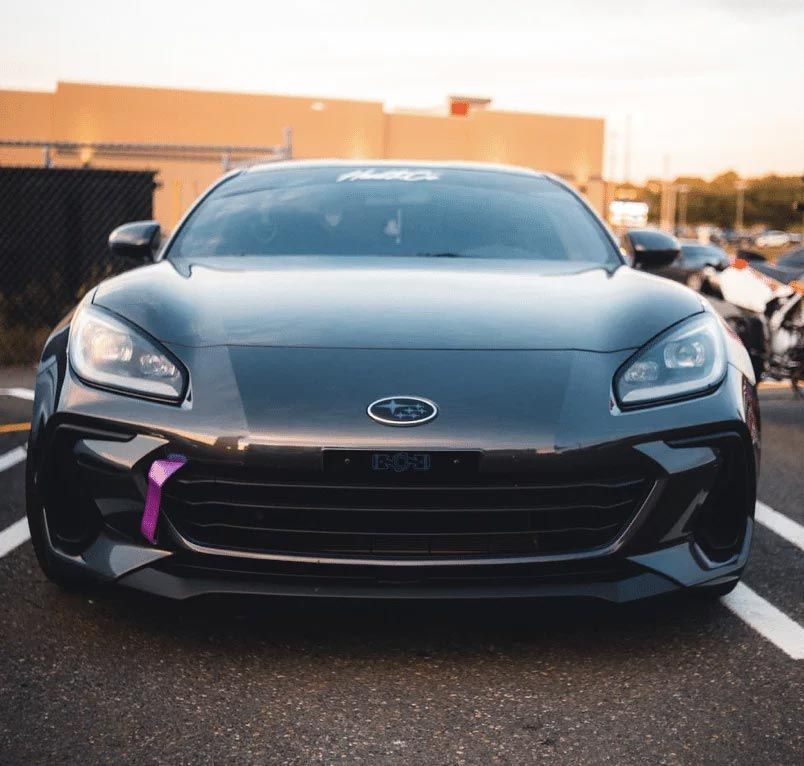 Black sports car parked front-facing outdoors at sunset, with a purple tow strap on the left side of the bumper