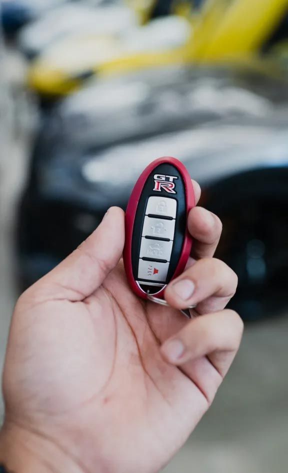 Hand holding a black and pink car key fob with three buttons, blurred yellow car in background