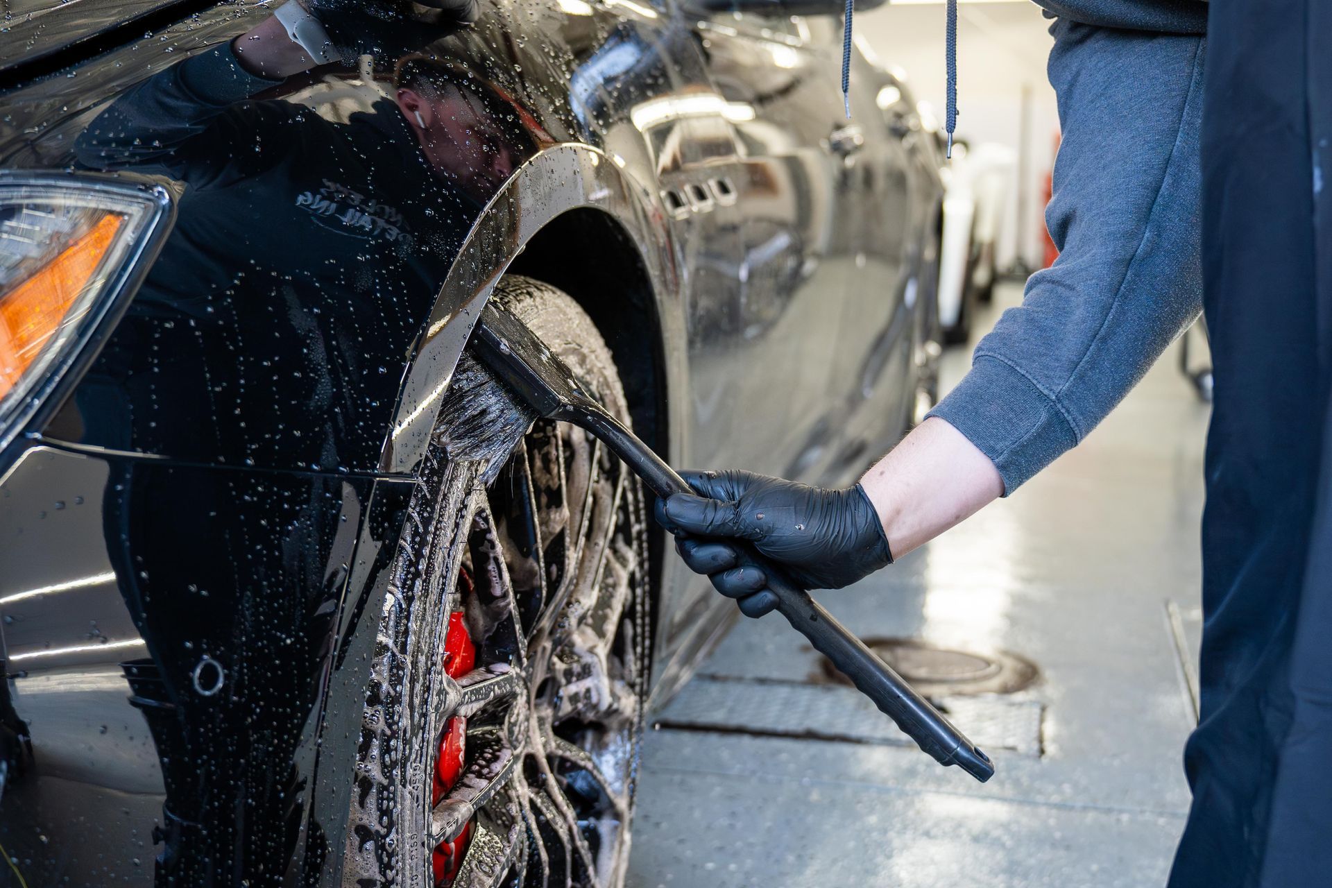 Person pressure-washes a black car wheel and fender outdoors on a wet driveway.