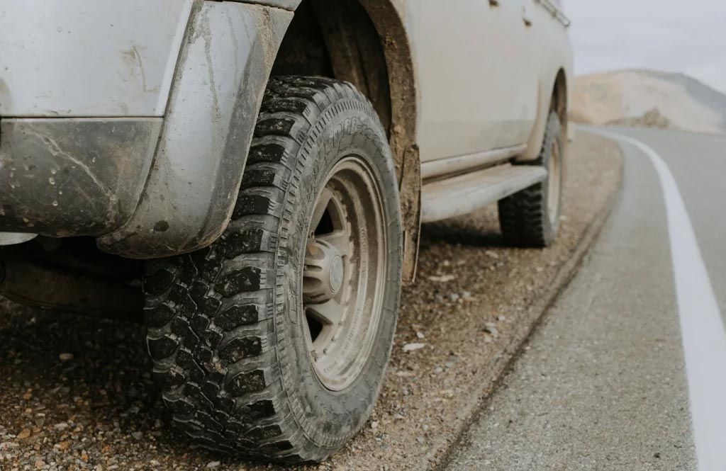 Close-up of a muddy pickup truck tire on a gravel shoulder beside a wet road