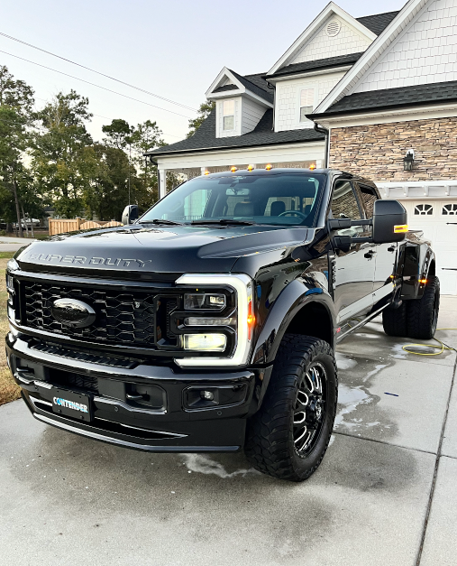 Black lifted Ford pickup truck parked in a driveway beside a house, with a trailer hitched behind it