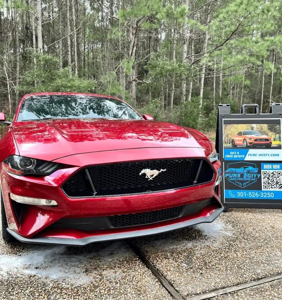 Red Ford Mustang parked beside a real estate sign on a driveway near trees