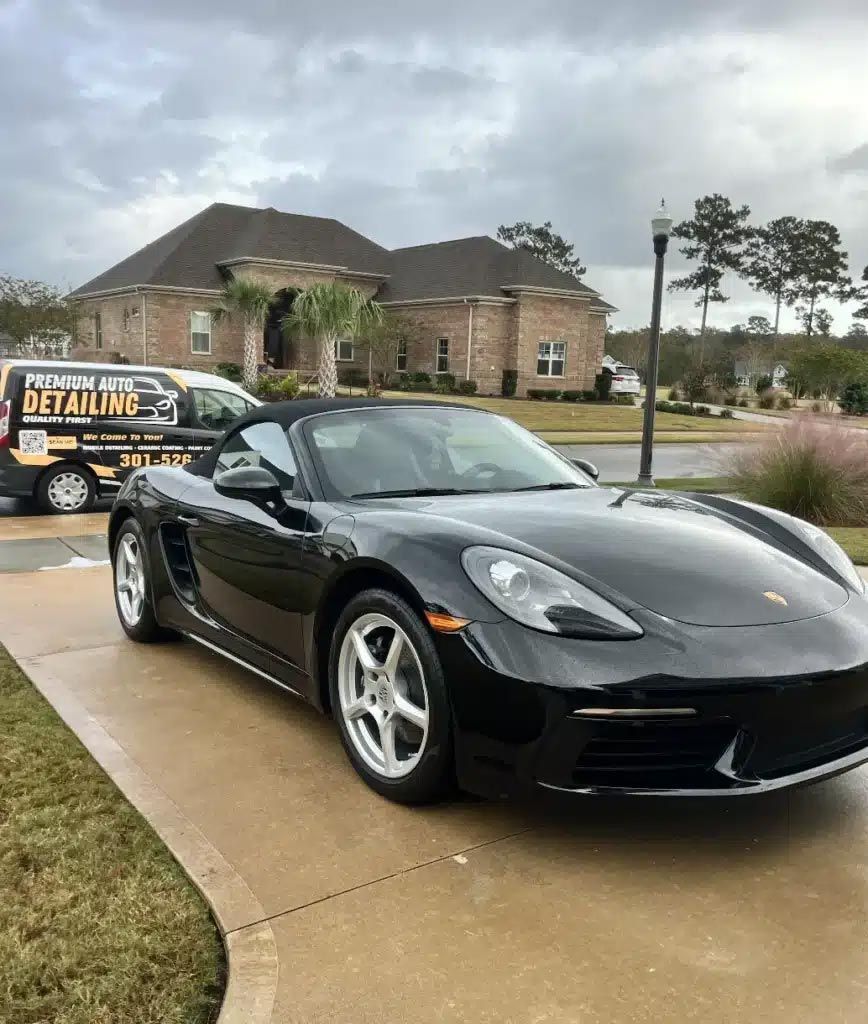 Black sports car parked in a driveway outside a brick house, with a detailing van in the background.