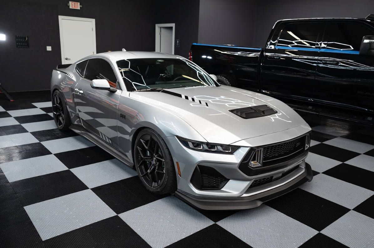 Silver sports car in a checkered-floor showroom, parked beside a black vehicle and a large truck.