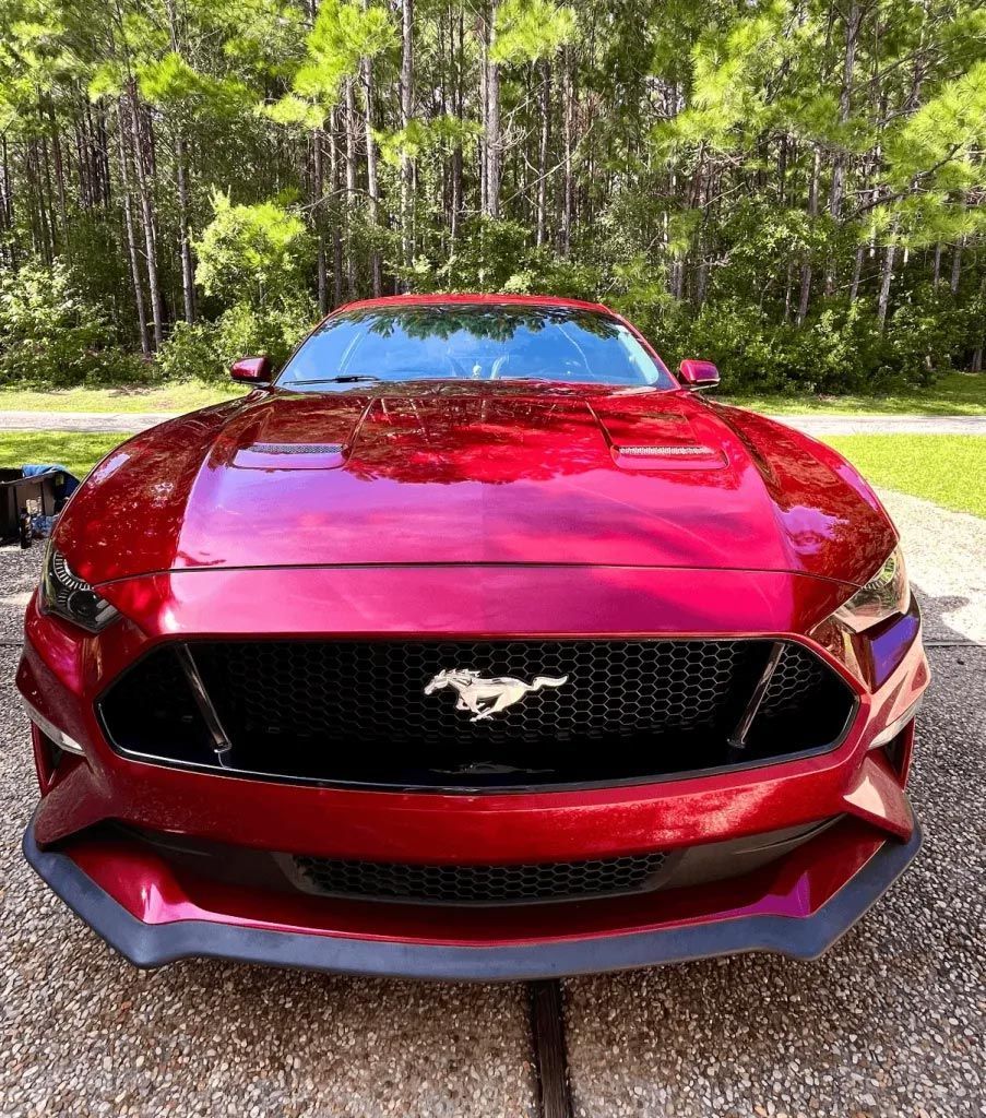 Red Ford Mustang parked on a gravel driveway with trees in the background.