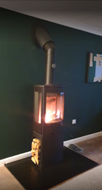 Modern wood burning stove with visible flame against a dark green wall and log storage below
