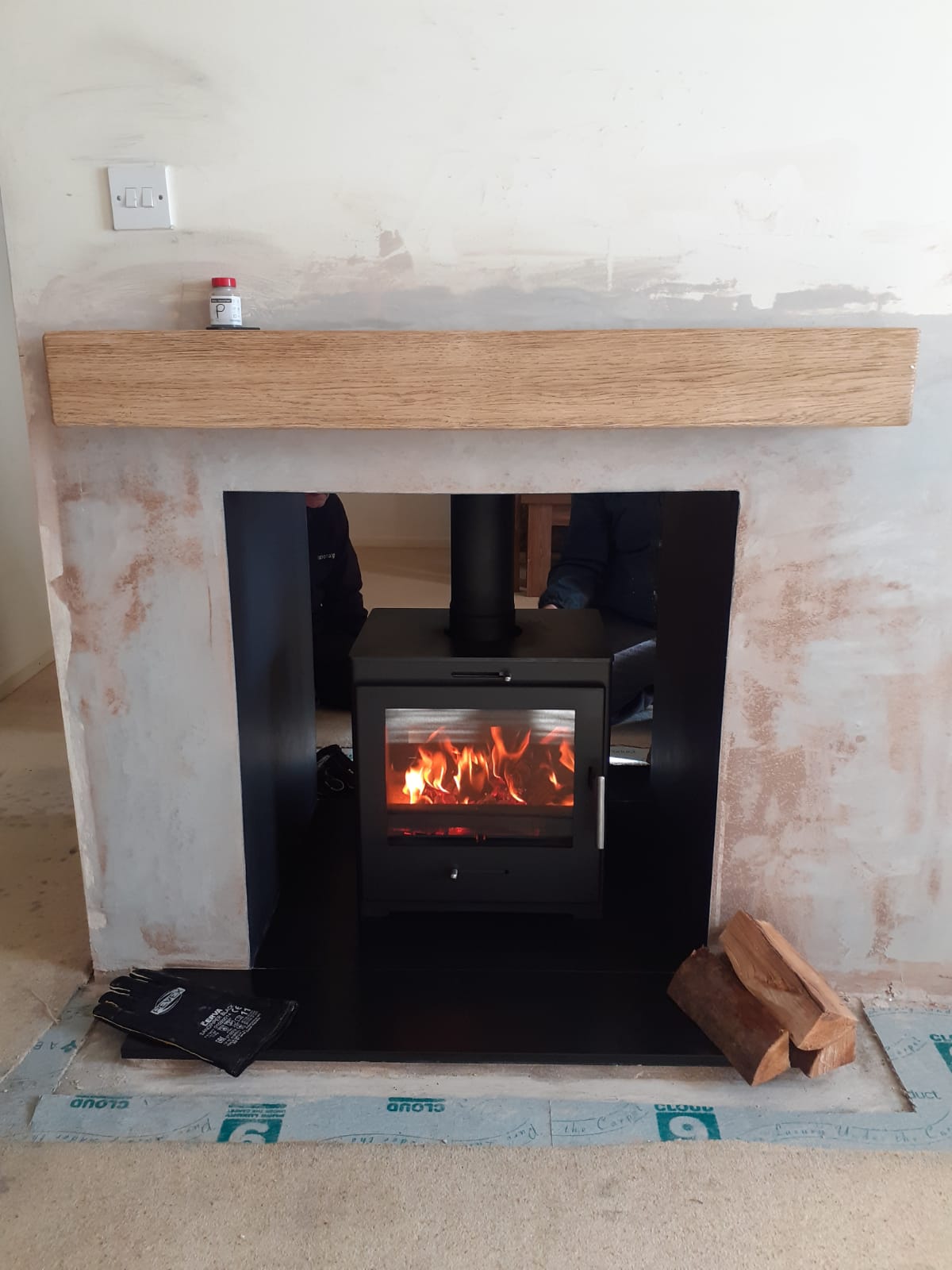 Wood burning stove with flame in a newly installed plastered fireplace and wooden mantel.