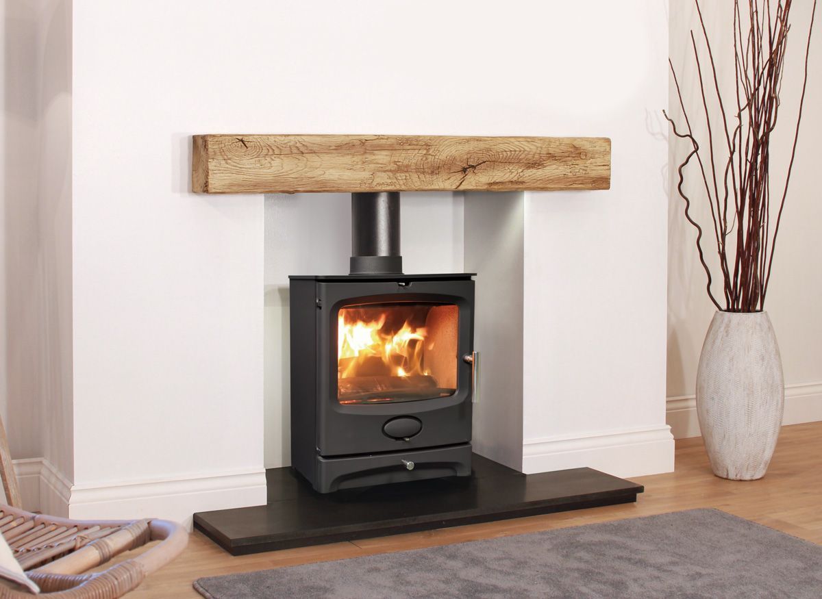 Black stove with flames in a white fireplace, under a wood beam mantle. Gray hearth, vase with reeds nearby.