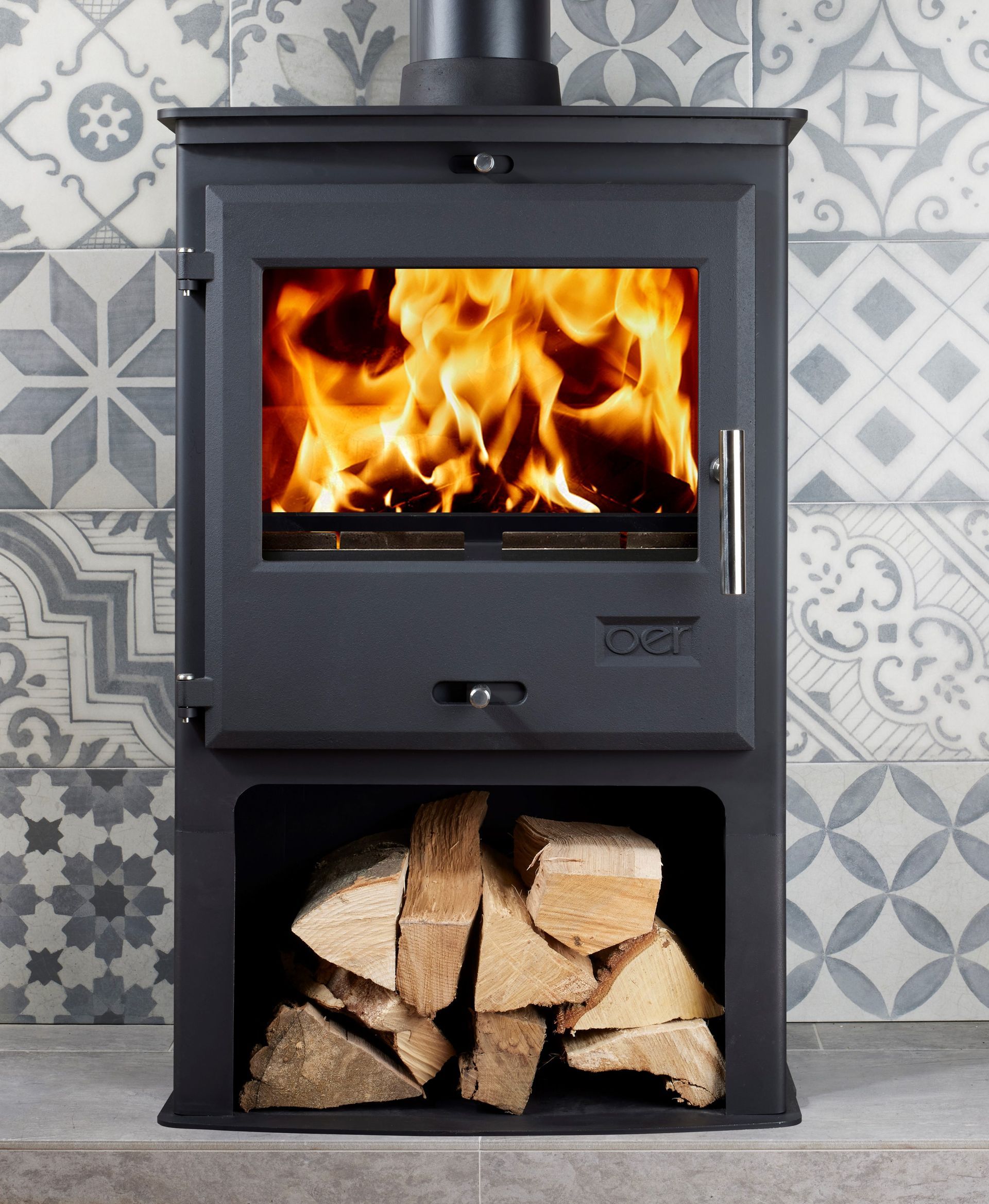 Black wood-burning stove with flames, firewood below, against patterned gray tile wall.
