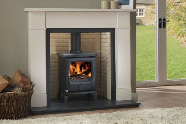 Wood-burning stove inside a cream-colored fireplace. Logs in a basket on the left. Flames inside the stove.