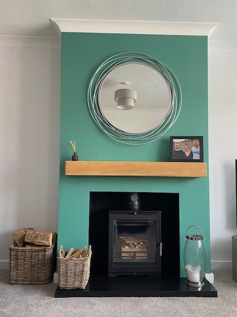 Wood burning stove with green accent wall, wooden shelf, and round mirror in a cosy living room