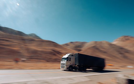 Un camión semirremolque negro circula por una carretera desértica con montañas borrosas al fondo.