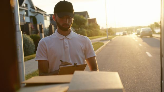 Un repartidor revisa la lista de paquetes junto a una calle soleada durante la hora dorada.