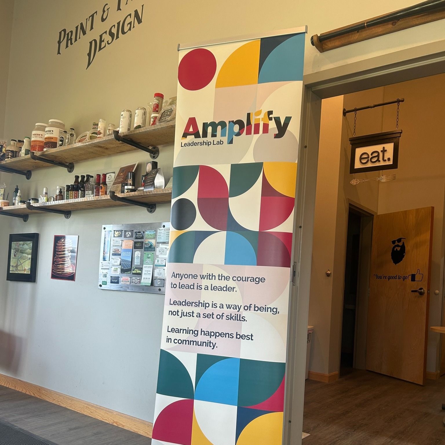 Banner for "Amplify" with colorful geometric design, near a door with "eat" sign. Shelves and wall text visible.