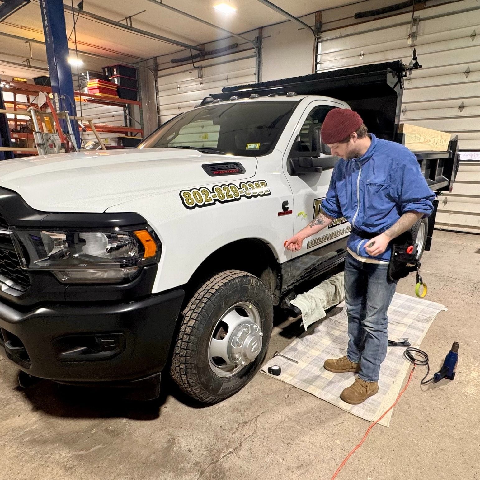 Man applying decal to white truck in a garage; uses tools and kneels on a mat.