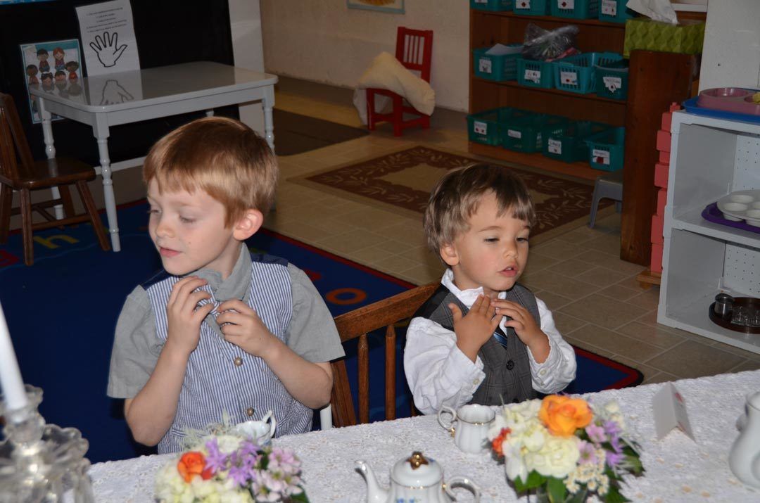 Two children in formal attire sit at a table with flower arrangements and tea service in a classroom setting.