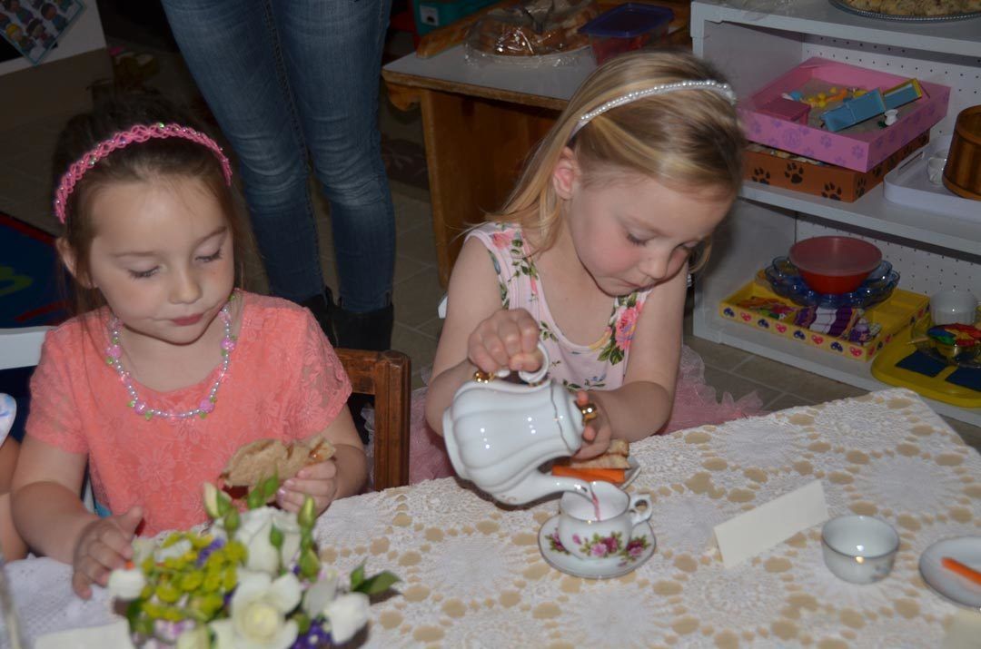 Two young children participate in a pretend tea party at a table set with a teapot, cups, and a small floral centerpiece.