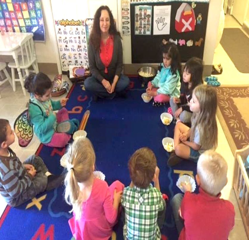 A teacher leads a group of children in a classroom activity, sitting in a circle on a blue rug with alphabet designs.