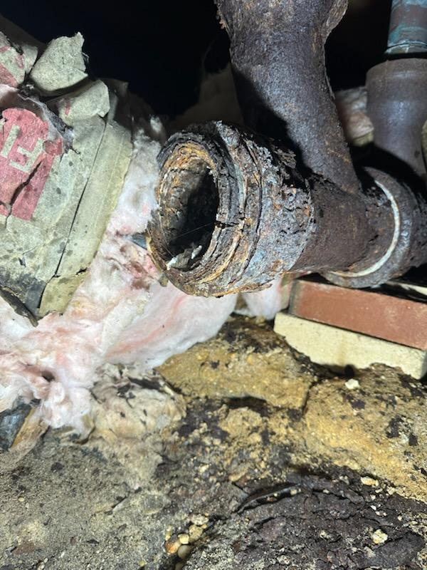 Severely corroded, rusted pipe joint. Brown rust, exposed inner components, surrounded by insulation, brick, and dirt.