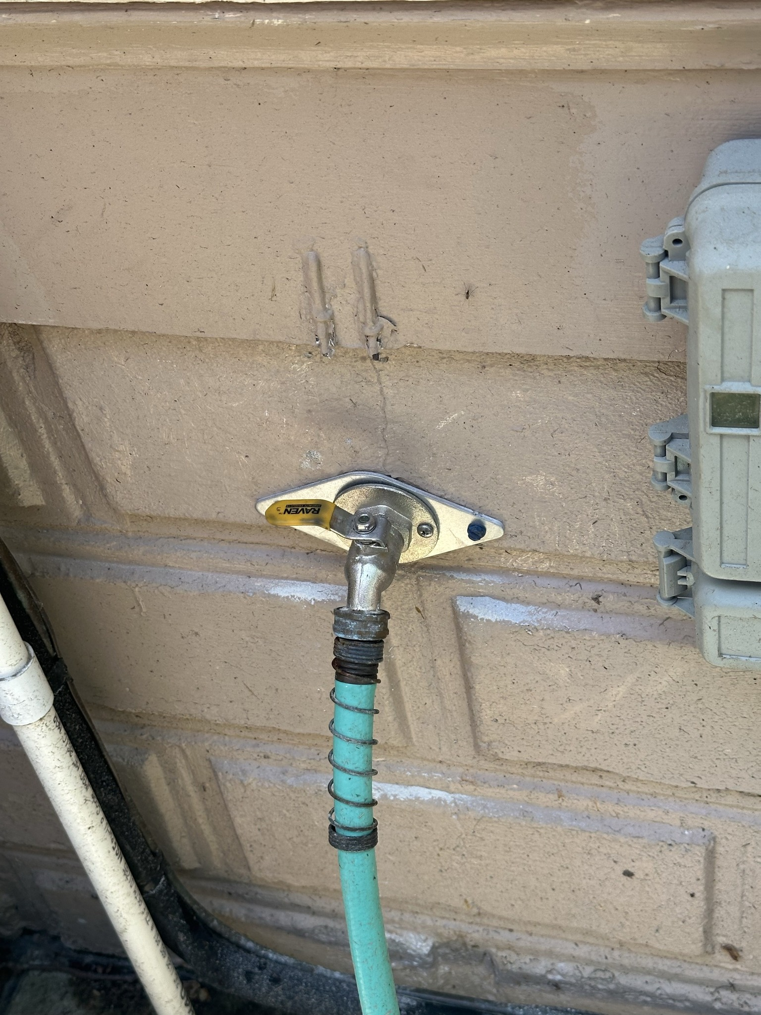 Outdoor spigot with green hose attached to a beige wall.  A white pipe and a white box are also present.