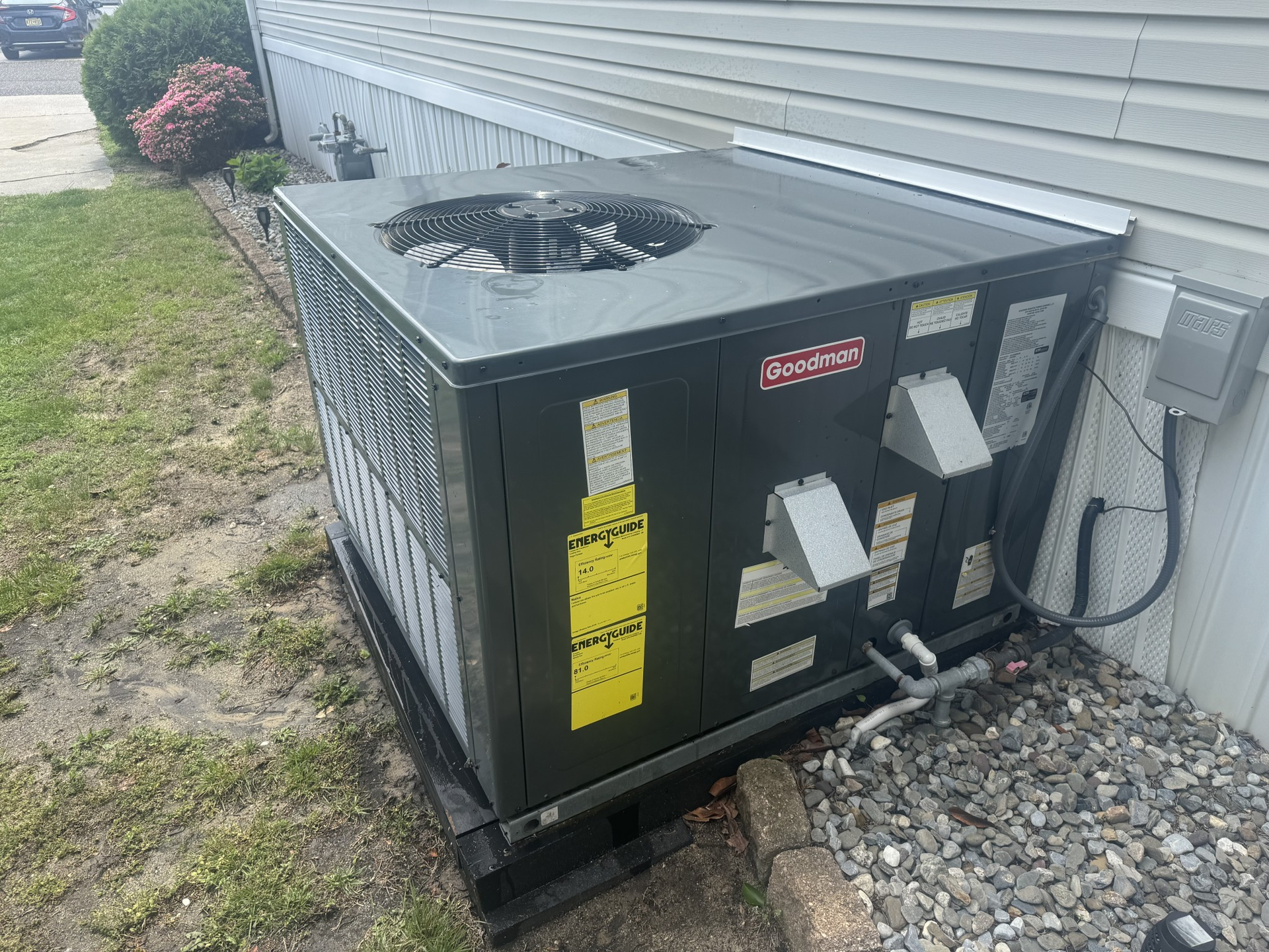 Black HVAC unit next to a light gray house on a gravel base in a yard with grass and bushes.