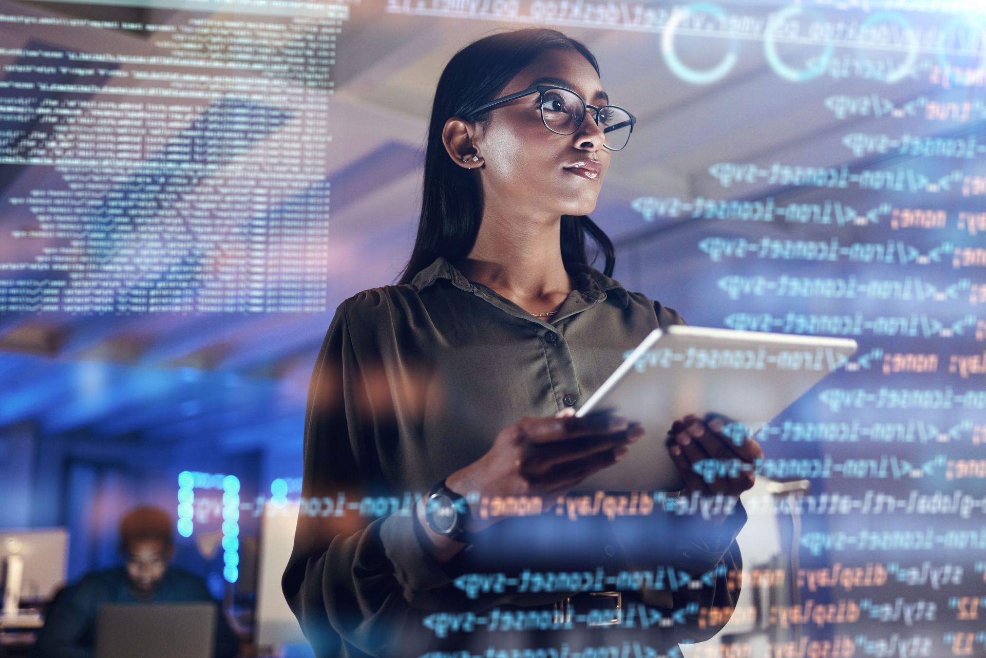Woman with glasses holding tablet, looking at projected code in a tech-filled office.