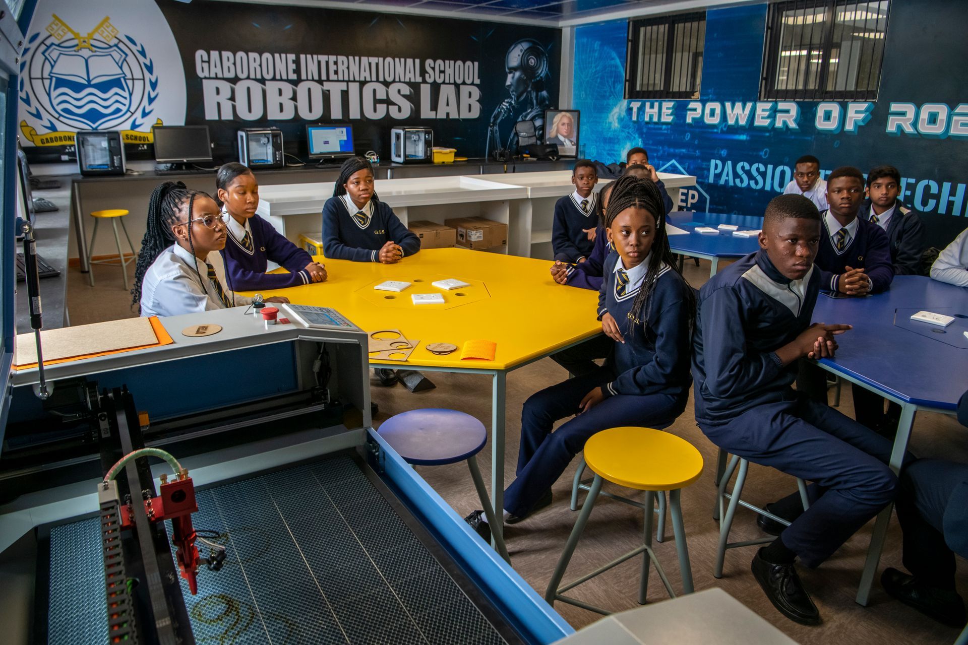 A group of children are sitting at tables in a robotics lab.