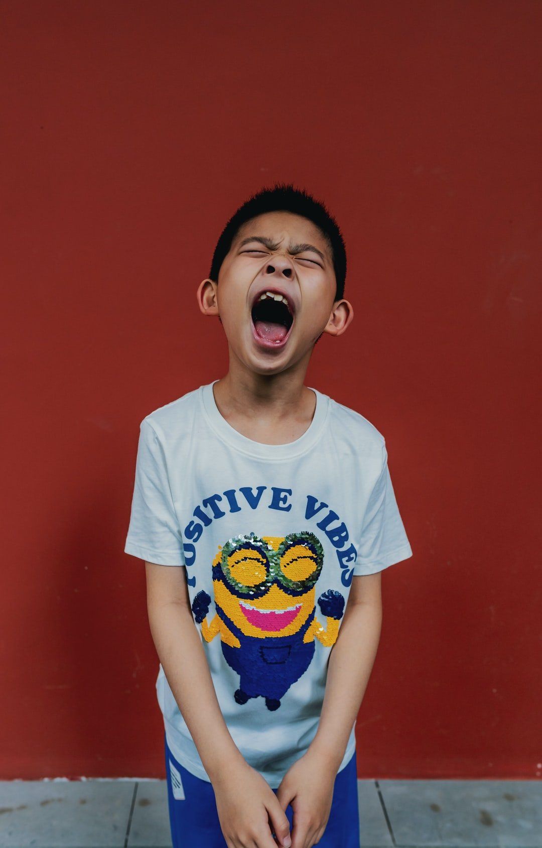 A young boy wearing a white t-shirt with a minion on it is standing in front of a red wall.