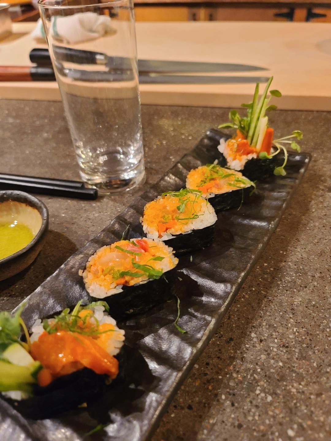 Sushi rolls on a black plate with orange topping, served beside a water glass and chopsticks at a restaurant