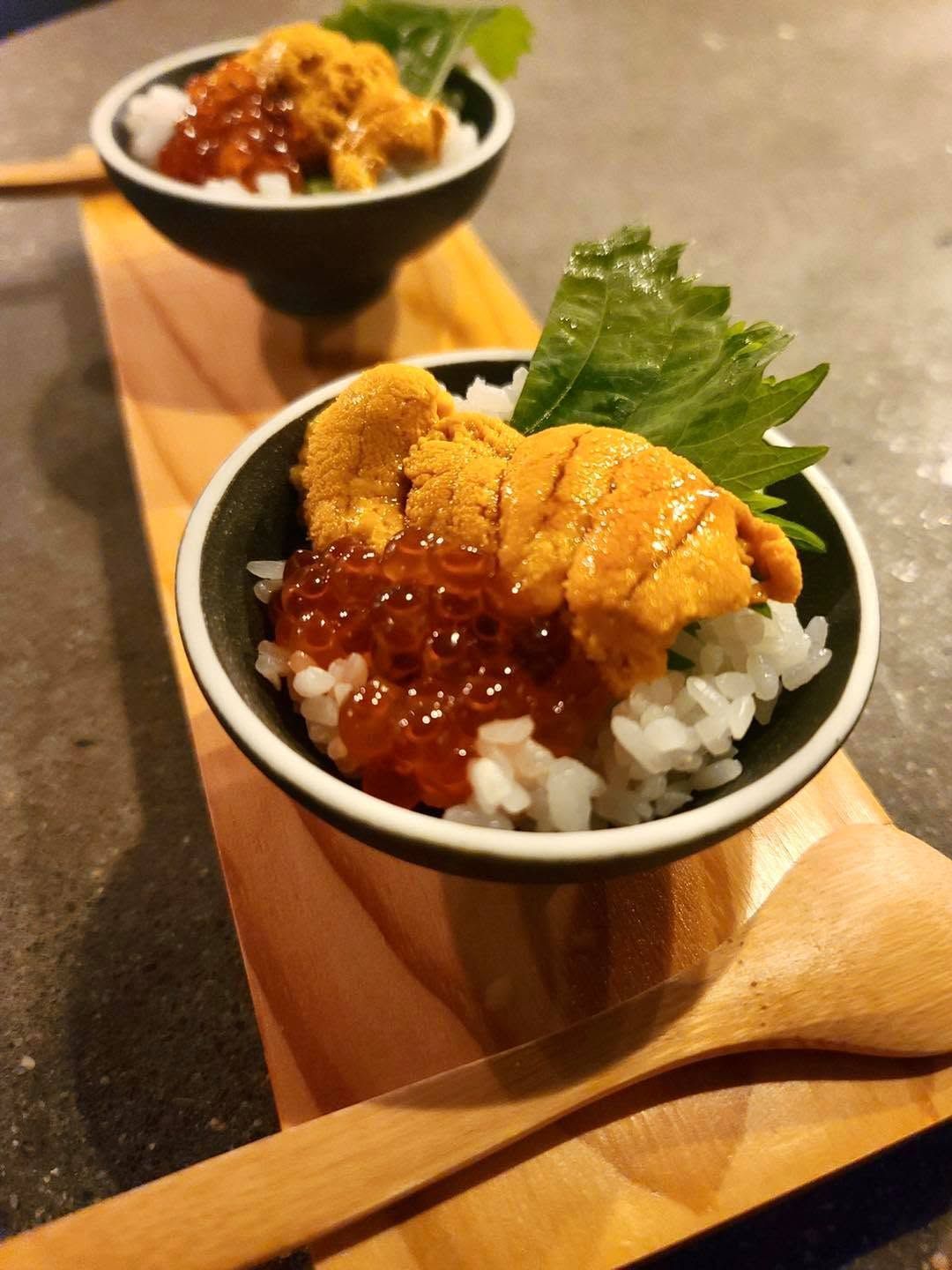 Two bowls of rice topped with sea urchin and sauce on wooden trays with spoons