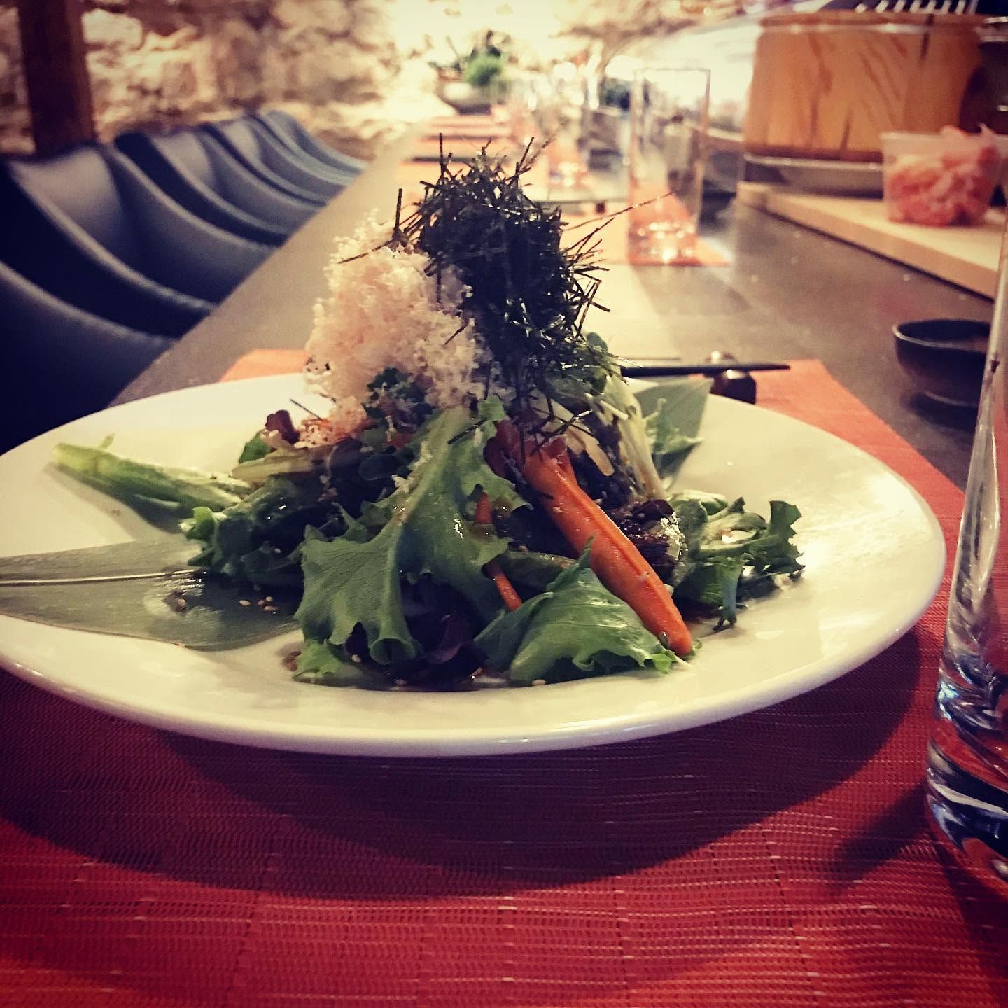 Salad on a white plate with greens and shredded garnish on a red tablecloth in a restaurant.