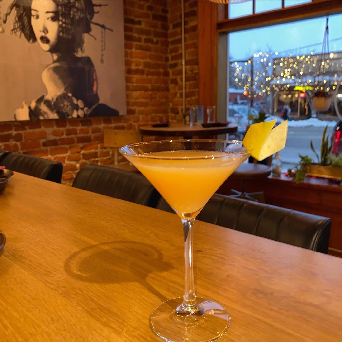 Martini glass with a bright orange cocktail and lemon garnish on a wooden bar in a cozy brick-walled café