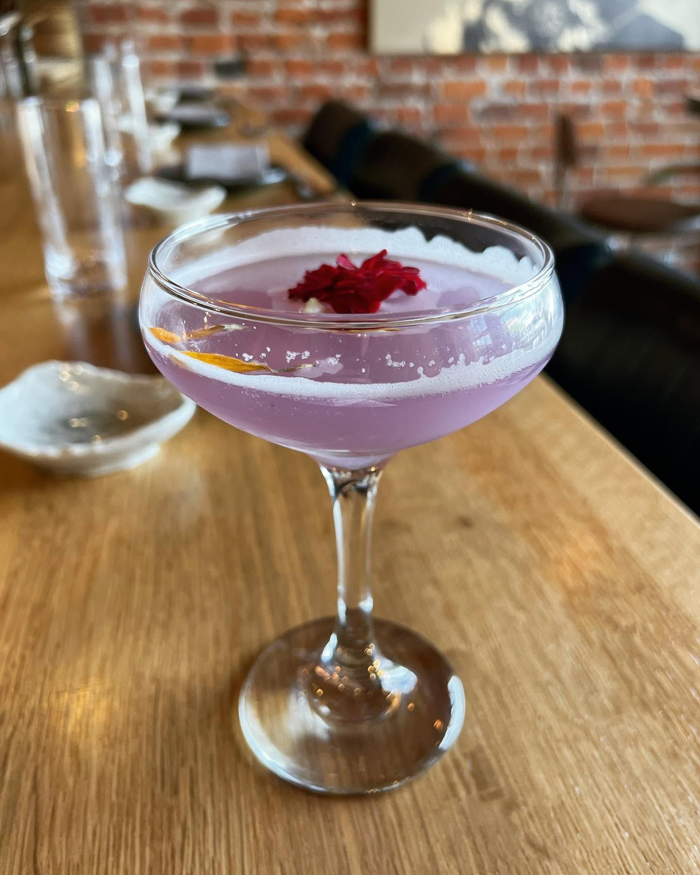 Lavender cocktail in a coupe glass with pink garnish on a wooden table in a brick-walled bar