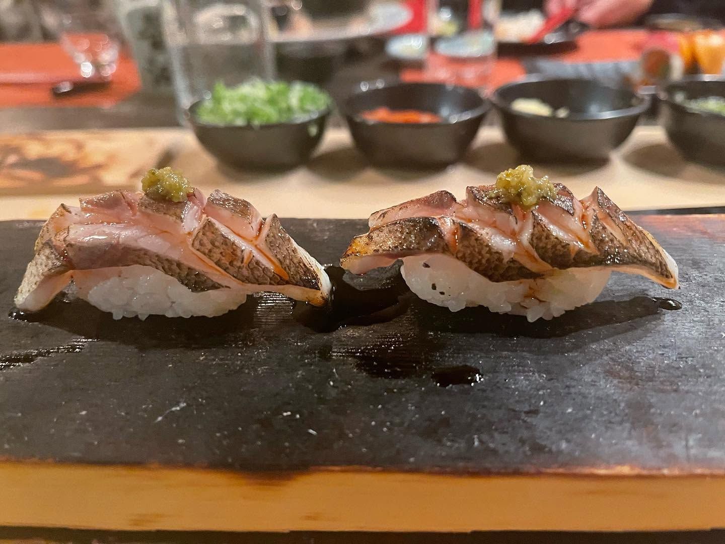 Two pieces of seared sushi on a black plate, with small bowls in the blurred background.