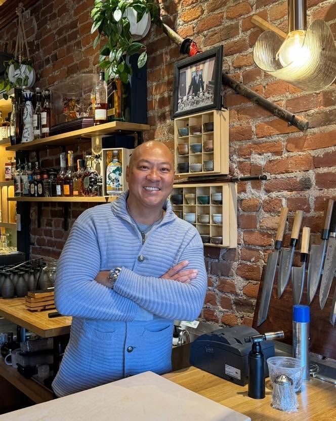 Smiling person in a blue sweater stands behind a bar with brick walls and liquor bottles.