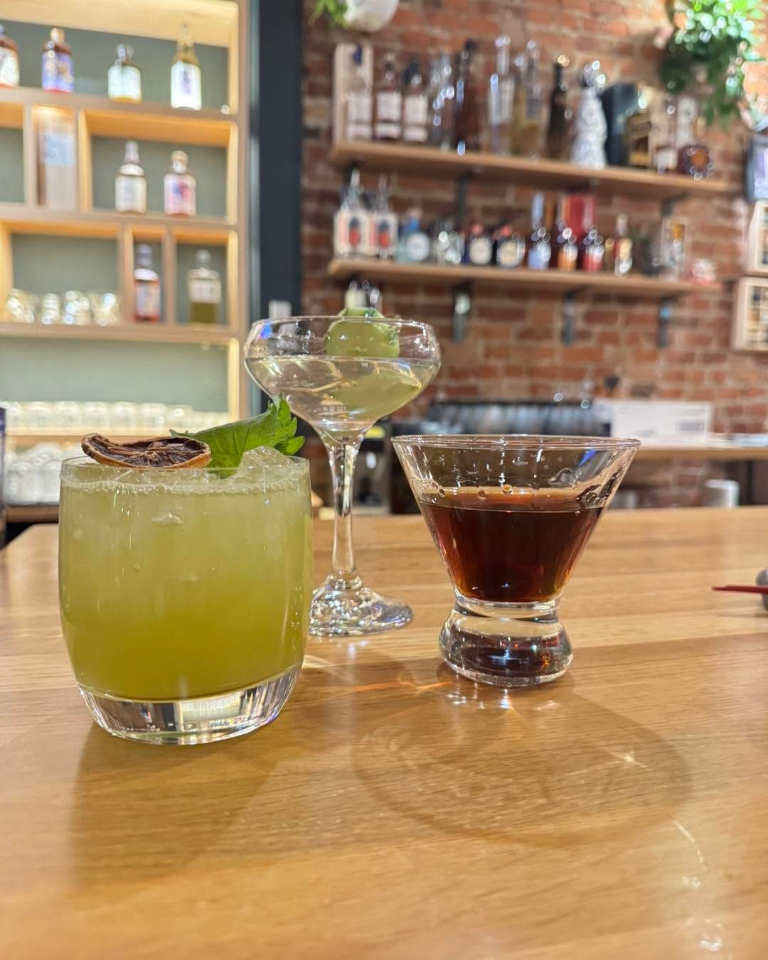 Three cocktails on a wooden bar, including a green drink, a martini, and a dark drink in a dim bar setting.