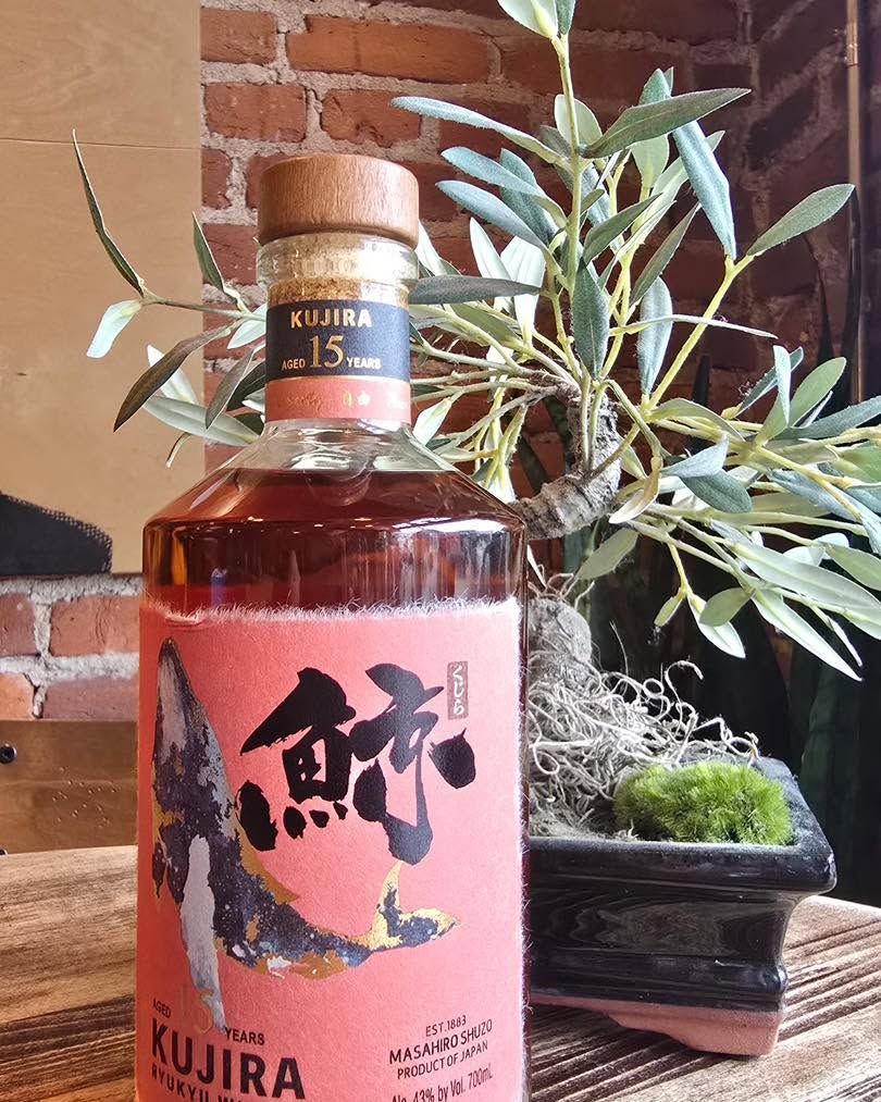 Bottle of Kujira whiskey with pink label on a table, beside a small potted plant.