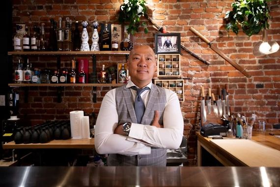 Bartender standing behind a bar with arms crossed in a brick-walled café, shelves and plants behind him.