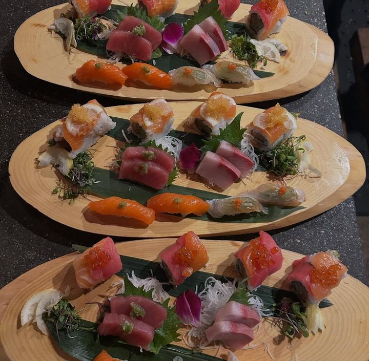 Three wooden platters of assorted sushi rolls and sashimi on a dark countertop