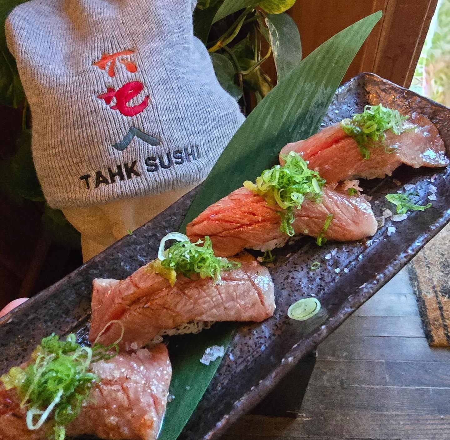 Sushi rolls topped with salmon and green garnish on a black plate, with a TAKK SUSHI bag in the background