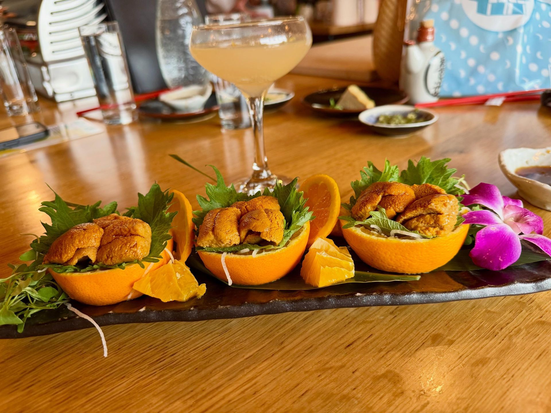 Three orange shell appetizers garnished with herbs on a dark platter, with drinks on a wooden table.