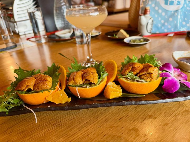 Three orange shell appetizers garnished with herbs on a dark platter, with drinks on a wooden table.