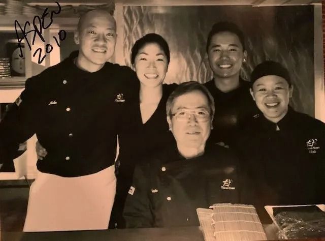 Five smiling people in dark shirts posing indoors behind a table with a cake, with a handwritten note in the corner