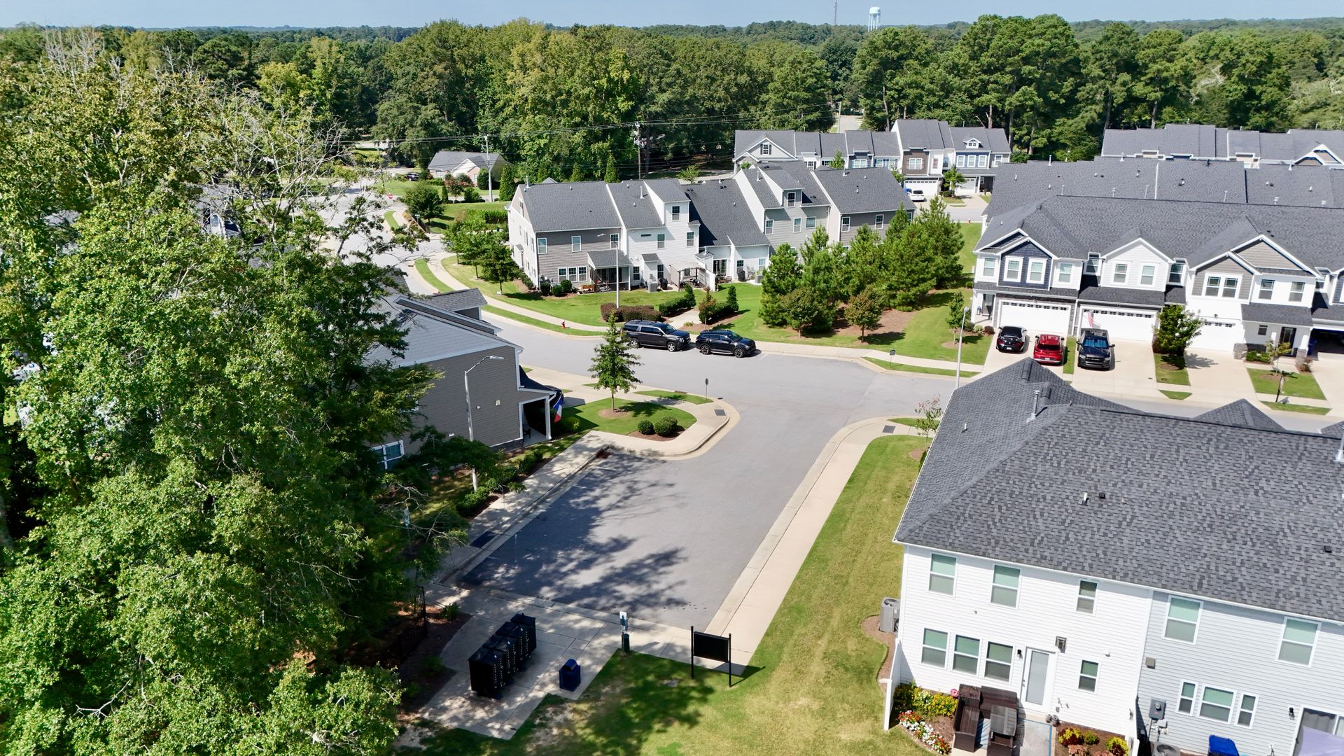Aerial view of a residential neighborhood with gray houses, green trees, and a street intersection under a blue sky.