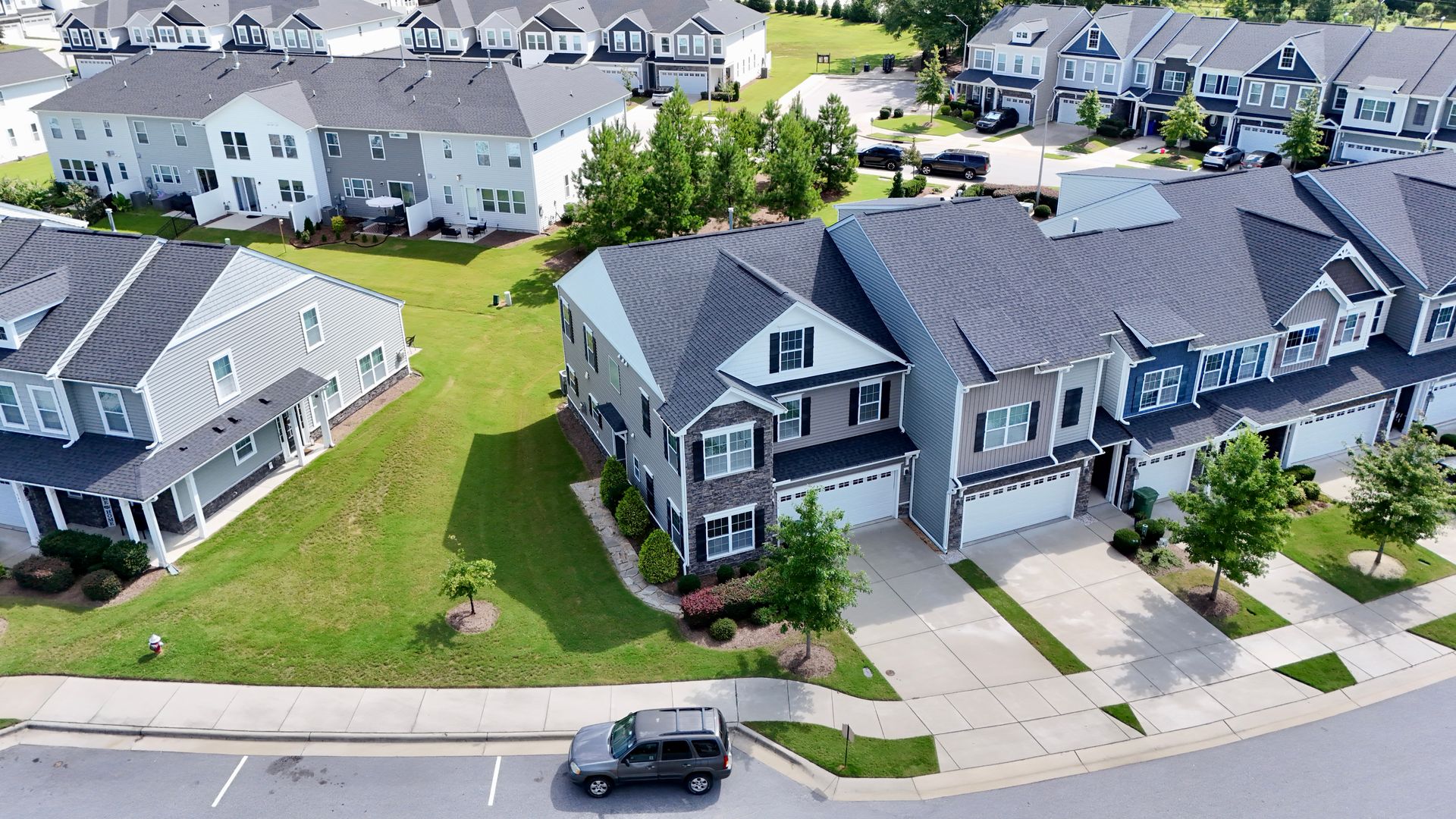 Aerial view of suburban houses with a car parked in front. Green lawns and blue sky.