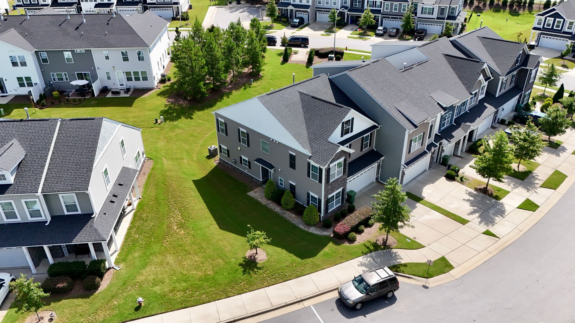 Aerial view of modern townhouses with gray and white exteriors, green lawns, and a parked SUV on a sunny day.