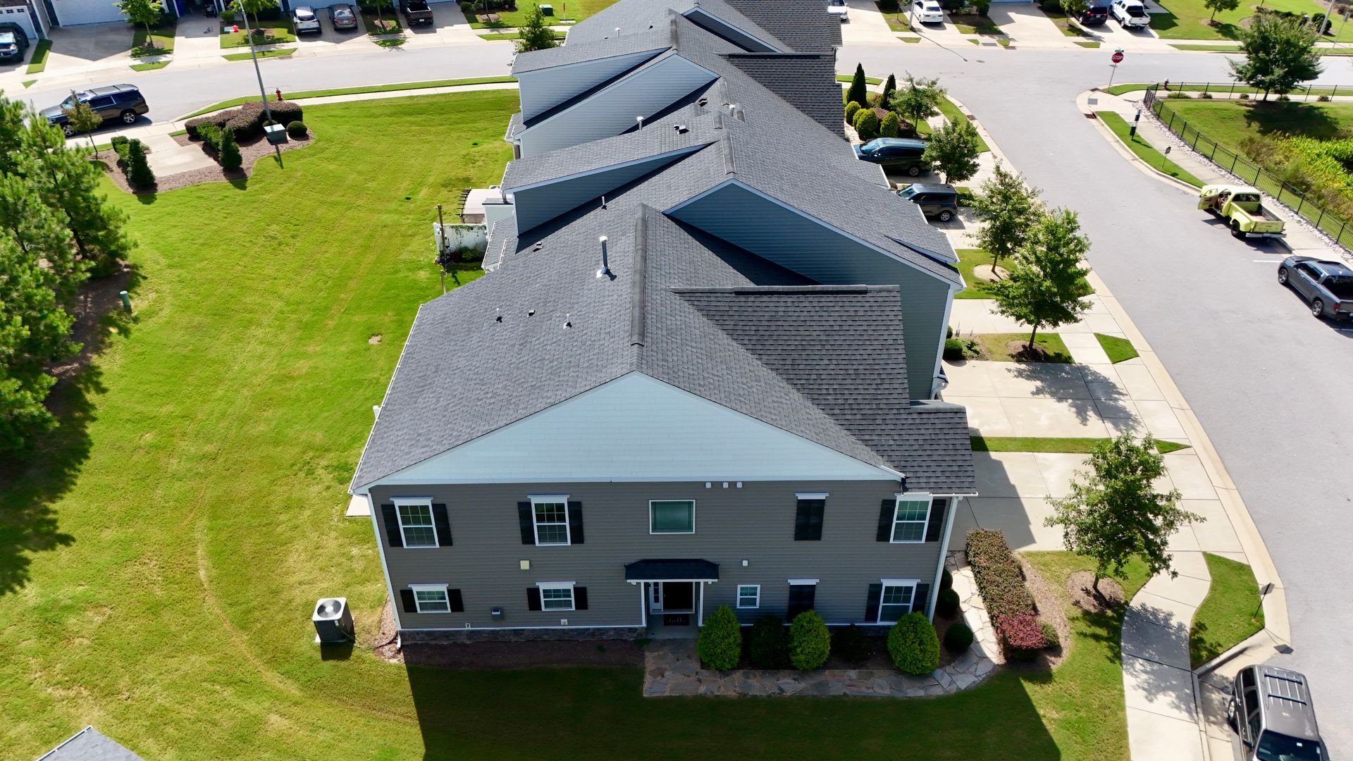 Aerial view of gray townhouses with dark roofs, green lawns, and street with cars.